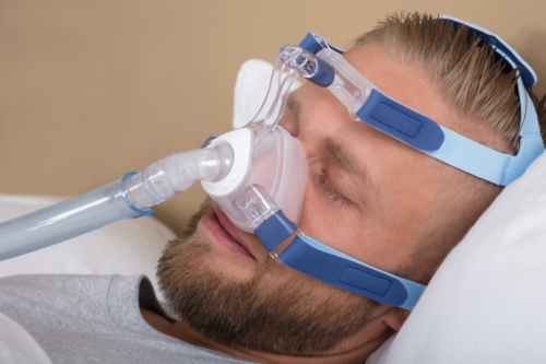 Young Man Lying On Bed With Sleeping Apnea And CPAP Machine