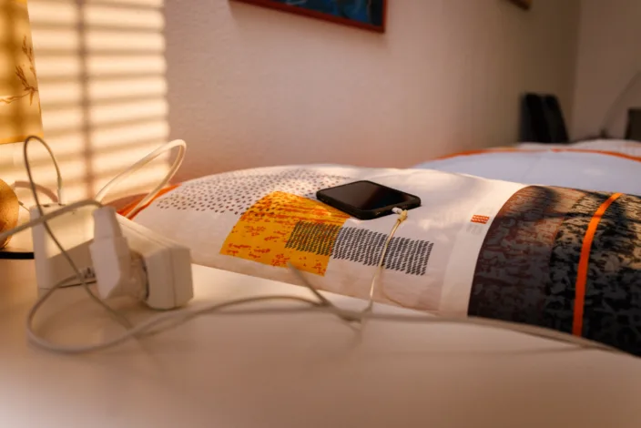 Ein Handy, eingesteckt zum aufladen, liegt auf dem Bett