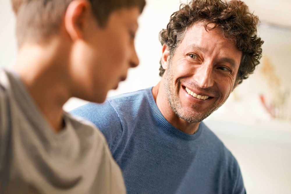 Nahaufnahme eines Vaters mit lockigem Haar, der seinen Sohn mit einem herzlichen, breiten Lächeln ansieht. Der Sohn im Vordergrund ist leicht unscharf, was den Fokus ganz auf den liebevollen und bejahenden Gesichtsausdruck des Vaters lenkt. Das Bild illustriert positive Vaterschaft, aktive Anteilnahme am Leben des Kindes und eine gelungene Kommunikation in der Familie. Es symbolisiert emotionale Sicherheit, die Stärkung des kindlichen Selbstwertgefühls durch väterliche Zuwendung und die Arbeit des Psychosozialen Gesundheitszentrums zur Förderung einer stabilen Vater-Kind-Bindung.