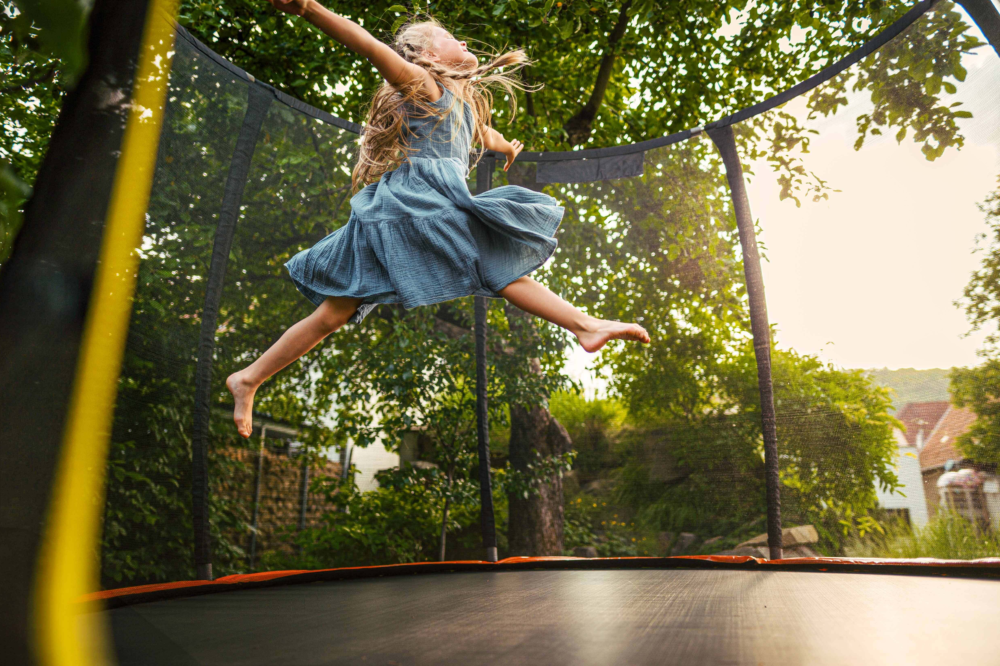 Ein junges Mädchen springt mit weit ausgebreiteten Armen und fliegenden Haaren voller Freude auf einem Gartentrampolin. Die Szene ist in warmes Sonnenlicht getaucht, im Hintergrund sind grüne Bäume zu sehen. Das Bild illustriert Bewegungsförderung bei Kindern, die Bedeutung von aktivem Spiel im Freien und pure Lebensfreude. Es symbolisiert den körperlichen Ausgleich zum digitalen Alltag, die Stärkung der Resilienz durch Sport und die ganzheitliche Begleitung des Psychosozialen Gesundheitszentrums für eine gesunde Kindheitsentwicklung.