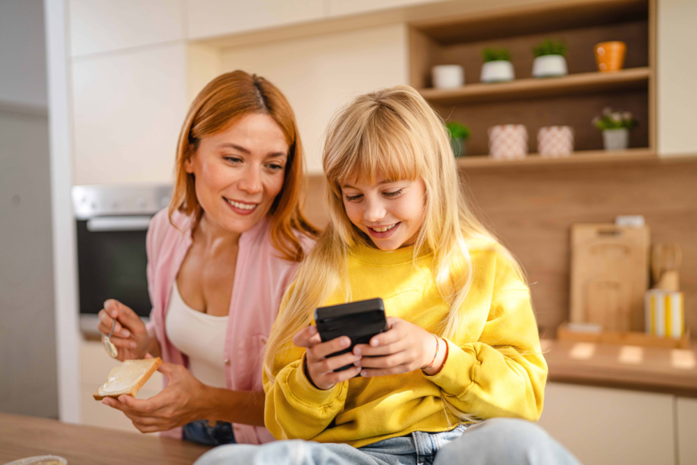 Eine Mutter und ihre Tochter in einer hellen, modernen Küche. Während die Mutter ein Brot streicht, blickt sie lächelnd und interessiert auf das Smartphone, das ihre Tochter stolz in den Händen hält. Das Bild illustriert die Integration von Medien in den Familienalltag, die Begleitung von Online-Aktivitäten durch Eltern und eine harmonische Kommunikation. Es symbolisiert das Vertrauensverhältnis bei der Handynutzung, die Vorbildfunktion der Eltern und die Beratung des Psychosozialen Gesundheitszentrums zur Förderung einer gesunden Medienkompetenz in der Familie.