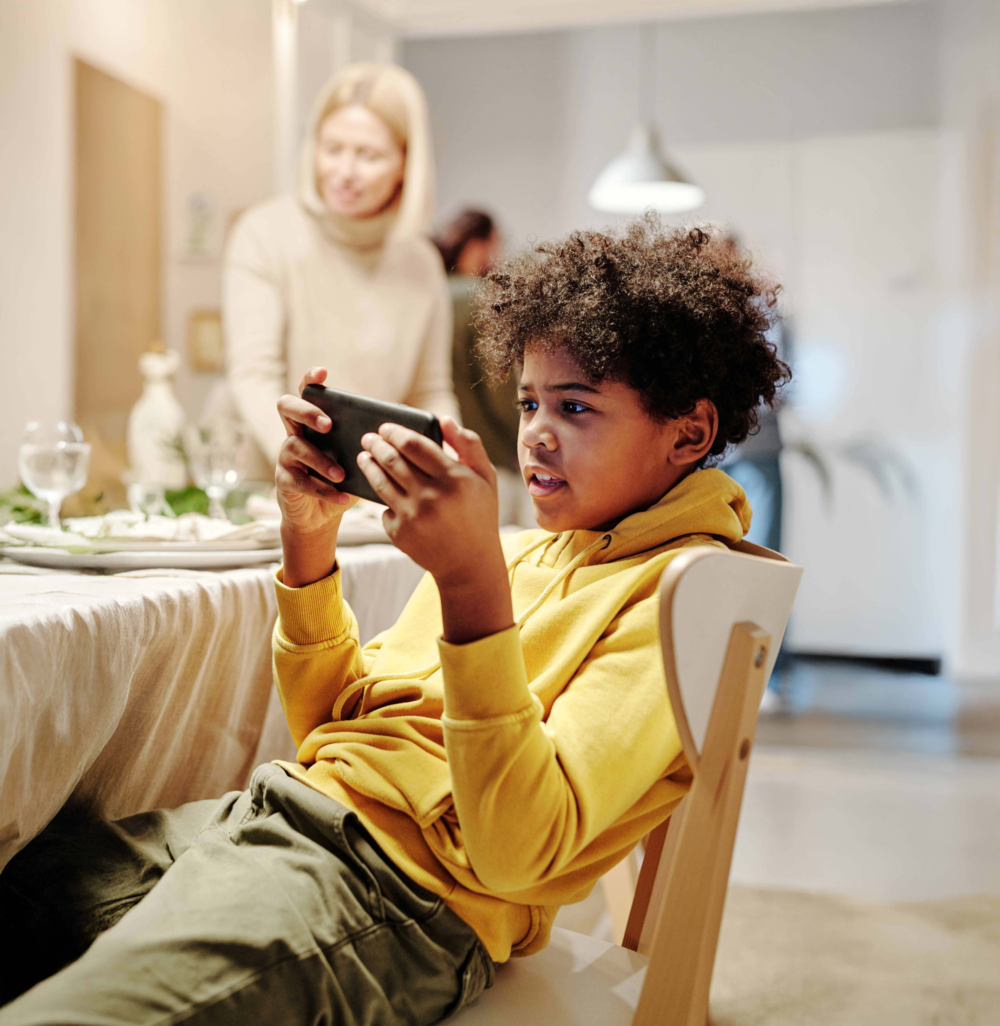 Ein Junge sitzt im Vordergrund auf einem Stuhl an einem festlich gedeckten Esstisch und spielt konzentriert auf einem Smartphone im Querformat. Im unscharfen Hintergrund ist eine Frau zu sehen, die den Tisch vorbereitet oder abräumt. Das Bild illustriert die soziale Isolation durch Medien während Familienmahlzeiten, die fehlende Interaktion am Esstisch und die Dominanz digitaler Spiele im Alltag. Es symbolisiert die Notwendigkeit von festen medienfreien Zeiten, die Beratung des Psychosozialen Gesundheitszentrums zu Erziehungskonflikten und die Förderung echter familiärer Begegnungen.