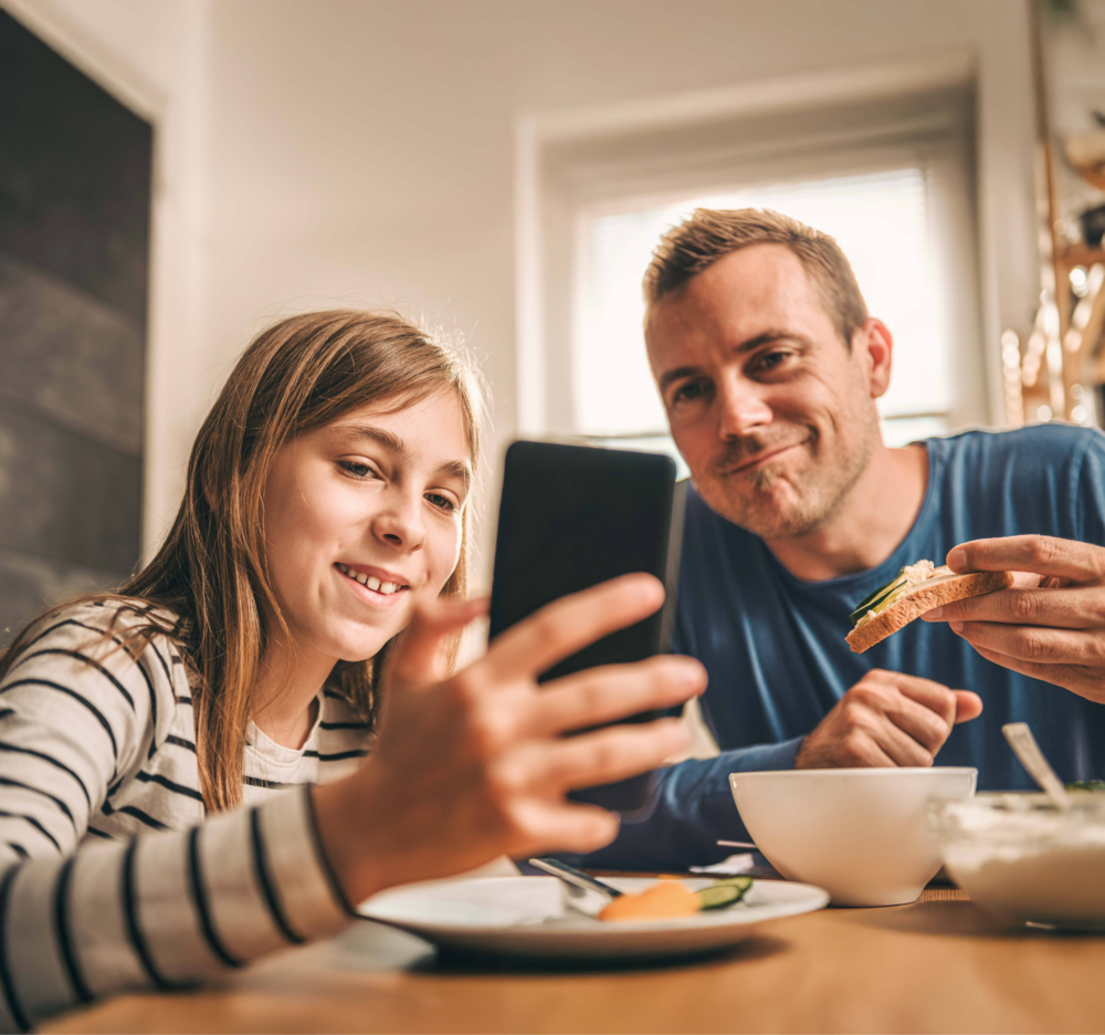 Ein Vater und seine Tochter sitzen gemeinsam am Frühstückstisch. Während sie essen, hält das Mädchen ein Smartphone in der Hand und zeigt dem Vater lächelnd etwas auf dem Display. Der Vater blickt entspannt und interessiert mit. Das Bild illustriert gemeinsame Mediennutzung, den lockeren Austausch über digitale Inhalte während der Mahlzeiten und eine positive Vater-Kind-Interaktion. Es symbolisiert die Begleitung der digitalen Welt durch Bezugspersonen und die offene Kommunikation über Online-Erlebnisse im geschützten familiären Rahmen.