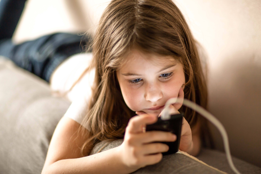 Nahaufnahme eines jungen Mädchens, das im Halbdunkel auf einem Sofa liegt und konzentriert auf ein Smartphone blickt, während das Gerät per Kabel aufgeladen wird. Das bläuliche Licht des Displays spiegelt sich in ihrem Gesicht wider. Das Bild illustriert übermäßigen Medienkonsum am Abend, die Herausforderung der digitalen Abgrenzung und das Phänomen des ‚Vampiring‘ (nächtliche Handynutzung). Es symbolisiert die Notwendigkeit von Schlafhygiene, Medienfasten und die Beratung des Psychosozialen Gesundheitszentrums bei beginnender Mediensucht oder Erschöpfung durch digitale Überreizung.