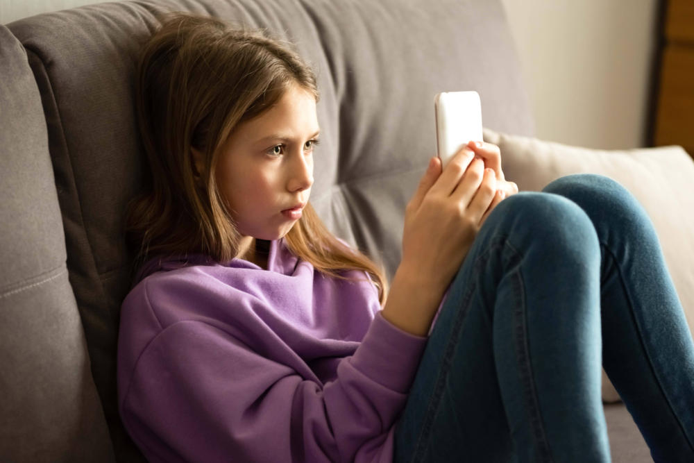 Ein Mädchen sitzt mit angezogenen Beinen in eine Sofaecke gedrückt und blickt mit einem sehr ernsten, fast bedrückten Gesichtsausdruck auf ihr Smartphone. Die Körperhaltung wirkt defensiv und isoliert. Das Bild illustriert die negativen Emotionen bei der Mediennutzung, die psychische Belastung durch soziale Netzwerke und die Anzeichen von Cybermobbing oder digitalem Stress. Es symbolisiert die Notwendigkeit von psychosozialer Beratung bei Internet-Sorgen, die Förderung von Resilienz im digitalen Raum und die Unterstützung des PSGZ für Jugendliche in emotionaler Not.