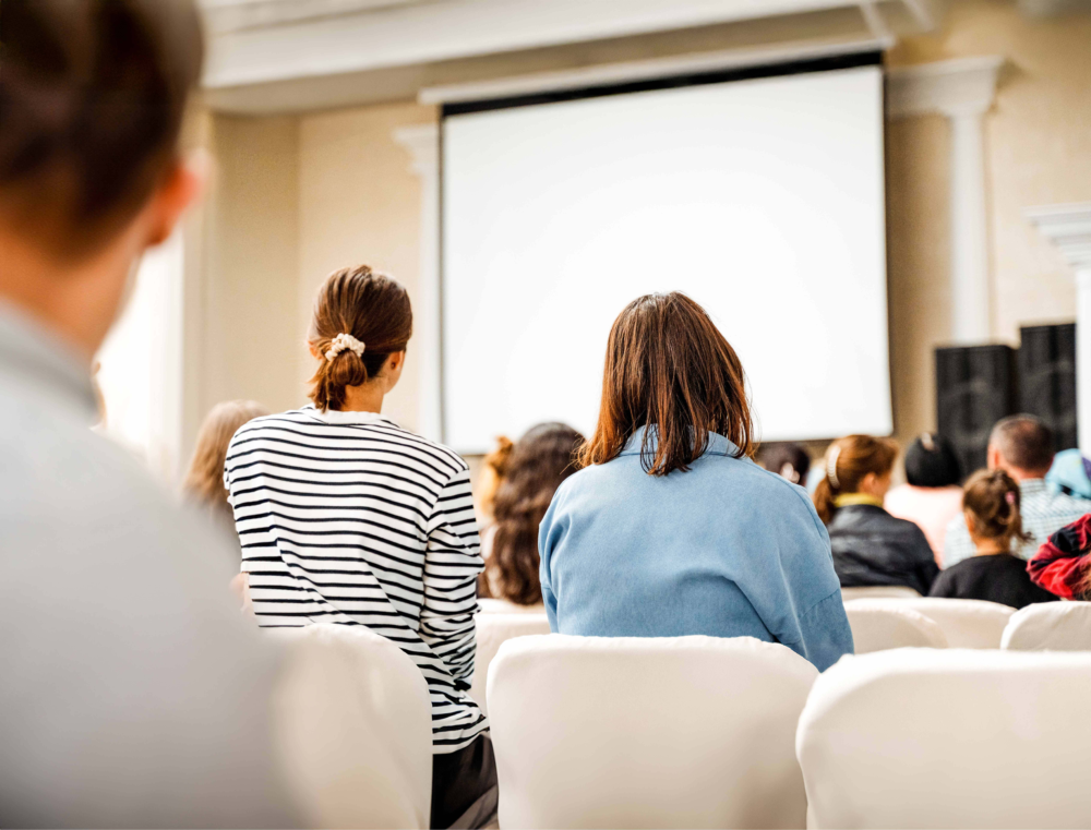 Blick aus dem Zuschauerraum auf ein Publikum, das gespannt auf eine große, weiße Projektionsfläche in einem Vortragssaal schaut. Die Szene illustriert eine Informationsveranstaltung, einen Fachvortrag oder einen thematischen Elternabend. Das Bild symbolisiert die Bildungsarbeit des Psychosozialen Gesundheitszentrums, den Austausch von Expertenwissen in der Gruppe und die Bedeutung von Prävention durch Aufklärung und öffentliche Veranstaltungen zu Erziehungs- und Gesundheitsthemen.