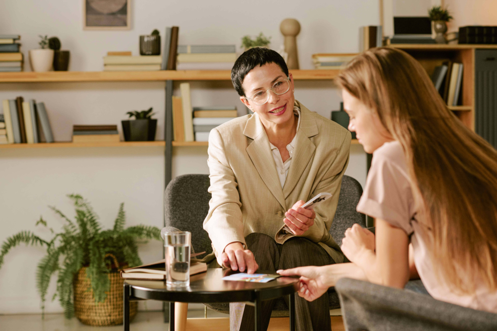 Eine erfahrene Beraterin sitzt einer jungen Frau in einem hellen, freundlich eingerichteten Praxisraum gegenüber. Die Beraterin hält eine Karte oder ein Dokument in der Hand und blickt ihr Gegenüber empathisch und unterstützend an. Das Bild illustriert eine professionelle Beratungssituation, die systemische Therapie oder ein Coaching-Gespräch. Es symbolisiert den sicheren Rahmen für persönliche Anliegen, professionelle Lebenshilfe und die lösungsorientierte Begleitung bei mentalen Herausforderungen durch das Psychosoziale Gesundheitszentrum.