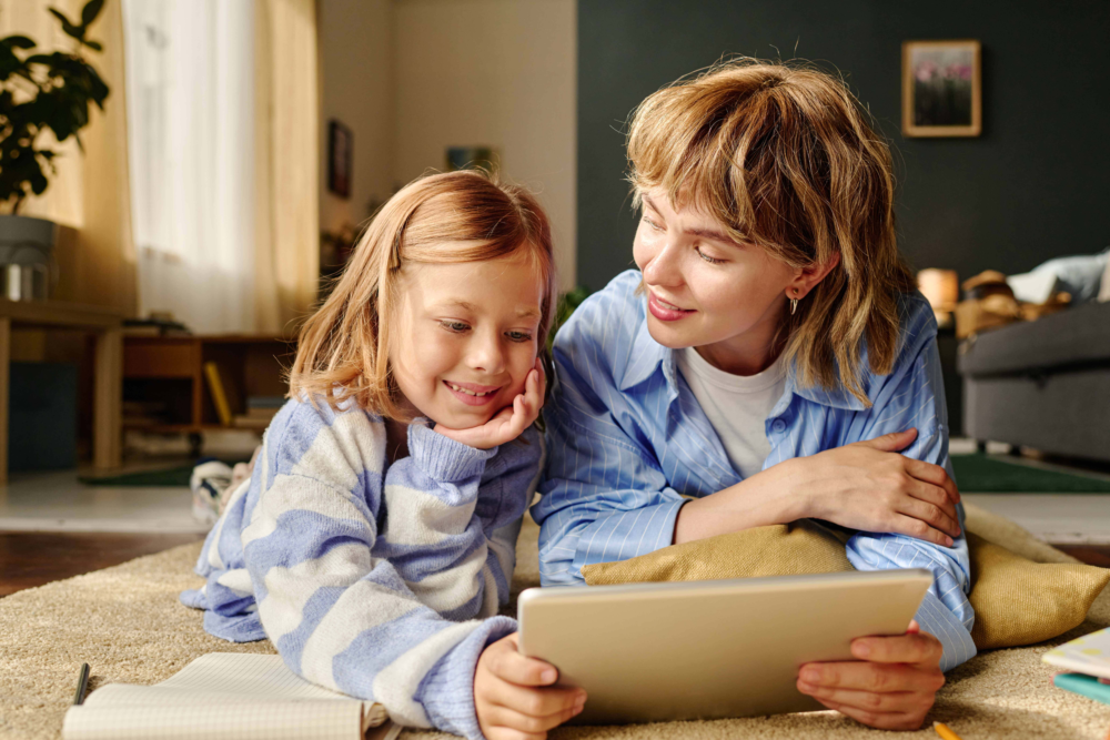 Eine Mutter und ihre Tochter liegen gemeinsam auf dem Boden eines gemütlichen Wohnzimmers und betrachten konzentriert ein Tablet. Beide lächeln und wirken entspannt während der digitalen Lerneinheit. Das Bild illustriert positive Lernbegleitung, die Nutzung von Bildungsmedien im familiären Umfeld und eine starke Mutter-Kind-Bindung. Es symbolisiert moderne Wissensvermittlung, spielerisches Lernen und die Unterstützung durch das Psychosoziale Gesundheitszentrum bei der Gestaltung einer förderlichen Lernatmosphäre zu Hause.