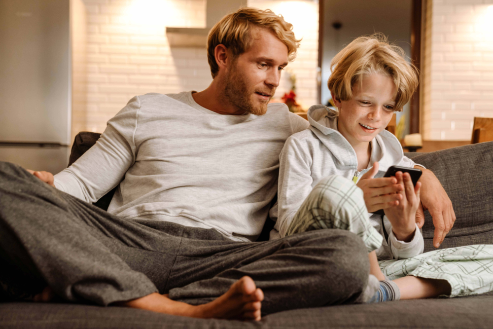 Ein Vater und sein Sohn sitzen entspannt nebeneinander auf einem dunklen Sofa. Der Junge hält ein Smartphone in den Händen und blickt lächelnd auf das Display, während der Vater aufmerksam und zugewandt mitschaut. Das Bild illustriert begleiteten Medienkonsum, das Interesse von Vätern an der digitalen Welt ihrer Kinder und eine harmonische Vater-Kind-Interaktion. Es symbolisiert die Förderung von Medienkompetenz durch gemeinsame Erlebnisse und einen sicheren Rahmen für die Smartphone-Nutzung zu Hause