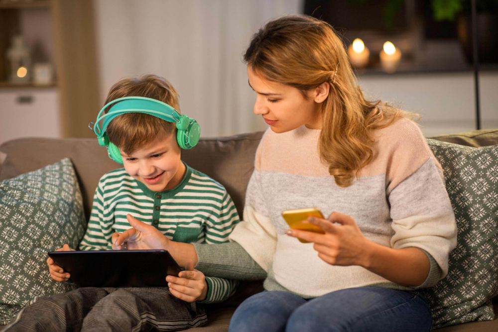 Eine Mutter und ihr Sohn sitzen gemeinsam auf einem Sofa und nutzen digitale Geräte. Der Junge trägt auffällige grüne Kopfhörer und bedient konzentriert ein Tablet, während die Mutter lächelnd neben ihm sitzt und ihr Smartphone hält. Das Bild illustriert begleiteten Medienkonsum, die Nutzung von Lern-Apps für Kinder und eine entspannte Atmosphäre bei der digitalen Wissensvermittlung. Es symbolisiert moderne Medienerziehung, die Förderung von IT-Kompetenz und die Unterstützung durch das Psychosoziale Gesundheitszentrum bei Fragen zur digitalen Balance.