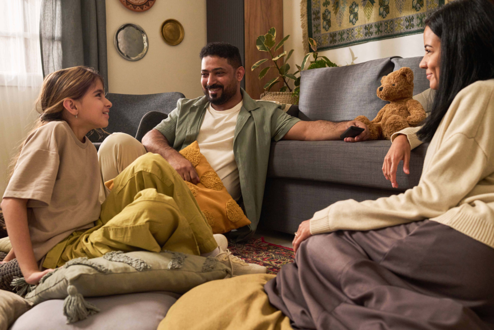 Eine Familie – bestehend aus Vater, Mutter und Tochter – sitzt entspannt und lachend gemeinsam auf dem Boden und Sofa in ihrem Wohnzimmer. Die Szene wirkt warm und einladend, mit weichen Kissen und einer gemütlichen Einrichtung. Das Bild illustriert positive Familiendynamik, den Wert von gemeinsamer Qualitätszeit und ein stabiles häusliches Umfeld. Es symbolisiert emotionale Sicherheit, soziale Integration und die präventive Arbeit des Psychosozialen Gesundheitszentrums zur Stärkung des Familienzusammenhalts.