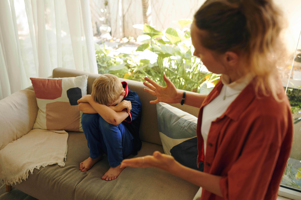 Ein Kind sitzt mit eingezogenen Knien und gesenktem Kopf auf einem Sofa und wirkt bedrückt, während im Vordergrund eine Frau mit ausladenden Handgesten spricht. Die Szene illustriert einen Erziehungskonflikt, Gefühle von Unverständnis und emotionale Belastung in der Familie. Es symbolisiert die Herausforderungen der Kommunikation zwischen Eltern und Kindern sowie den Bedarf an Strategien zur gewaltfreien Kommunikation und Deeskalation.