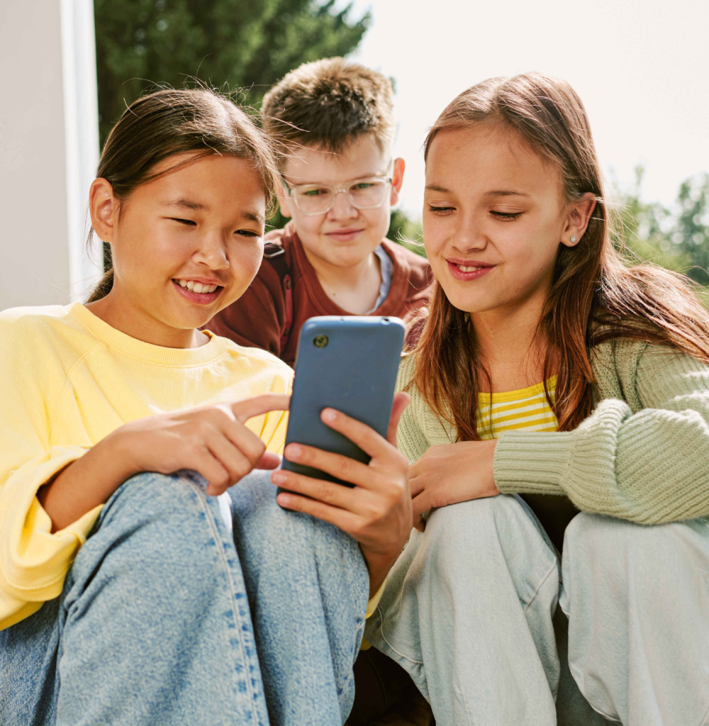 Drei Jugendliche sitzen gemeinsam im Freien und betrachten aufmerksam ein Smartphone. Sie lächeln und interagieren eng miteinander, während sie digitale Inhalte teilen. Das Bild illustriert die Bedeutung von sozialen Medien für Teenager, die gemeinschaftliche Mediennutzung und den Stellenwert von Peer-Interaktion im digitalen Zeitalter. Es symbolisiert Freundschaft, digitale Teilhabe und die Herausforderungen sowie Chancen der jugendlichen Medienwelt.