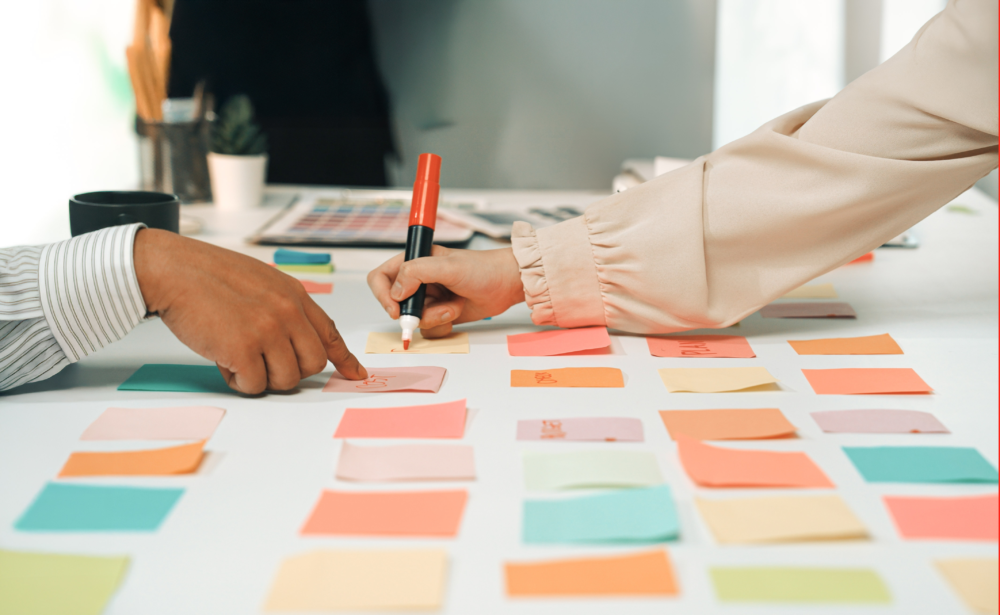 Nahaufnahme von zwei Personen, die an einem weißen Tisch zusammenarbeiten. Eine Person beschriftet mit einem roten Marker ein gelbes Klebenotiz-Blatt (Post-it), während eine andere Person auf ein benachbartes Blatt deutet. Der Tisch ist mit zahlreichen bunten Notizzetteln in einem Raster angeordnet. Das Bild illustriert agile Arbeitsmethoden, Design Thinking und die Visualisierung von Ideen. Es symbolisiert Teamwork, Projektmanagement und die strukturierte Lösungsfindung in kreativen oder geschäftlichen Prozessen.