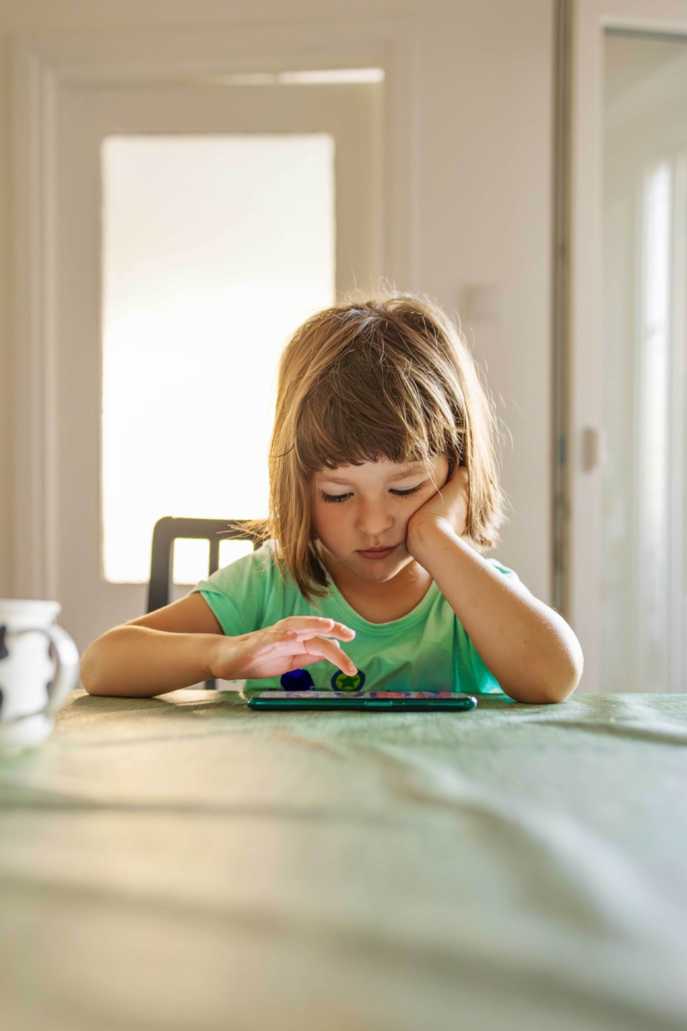 Ein kleines Kind sitzt allein an einem großen Esstisch und bedient mit gesenktem Kopf ein Smartphone, das flach auf der Tischdecke liegt. Das Kind stützt den Kopf auf eine Hand, während die andere Hand den Bildschirm berührt. Im Hintergrund ist eine helle Zimmertür zu sehen. Das Bild illustriert frühzeitige Handynutzung, die passive Körperhaltung bei digitalem Konsum und die Herausforderung, bildschirmfreie Zonen im Familienalltag zu etablieren. Es symbolisiert die Allgegenwärtigkeit von Technik, potenzielle Isolation am Esstisch und die Relevanz einer bewussten Begleitung der Medienzeit von Kleinkindern.