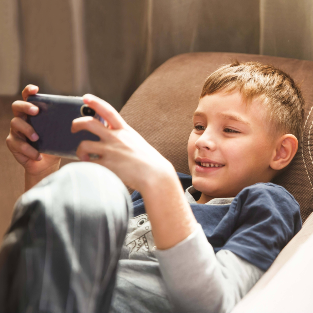 Nahaufnahme eines Jungen, der entspannt in einem Sessel sitzt und lächelnd ein Smartphone im Querformat hält. Er ist sichtlich in ein Video oder ein Spiel vertieft. Das Bild illustriert selbstbestimmten Medienkonsum bei Kindern, die Faszination digitaler Unterhaltung und die Bedeutung von Bildschirmzeit im modernen Kinderalltag. Es symbolisiert digitale Freizeitgestaltung, die Notwendigkeit von Medienkompetenz und die Beratung des Psychosozialen Gesundheitszentrums zur Begleitung von Kindern in der digitalen Welt sowie zur Balance zwischen Online- und Offline-Aktivitäten.