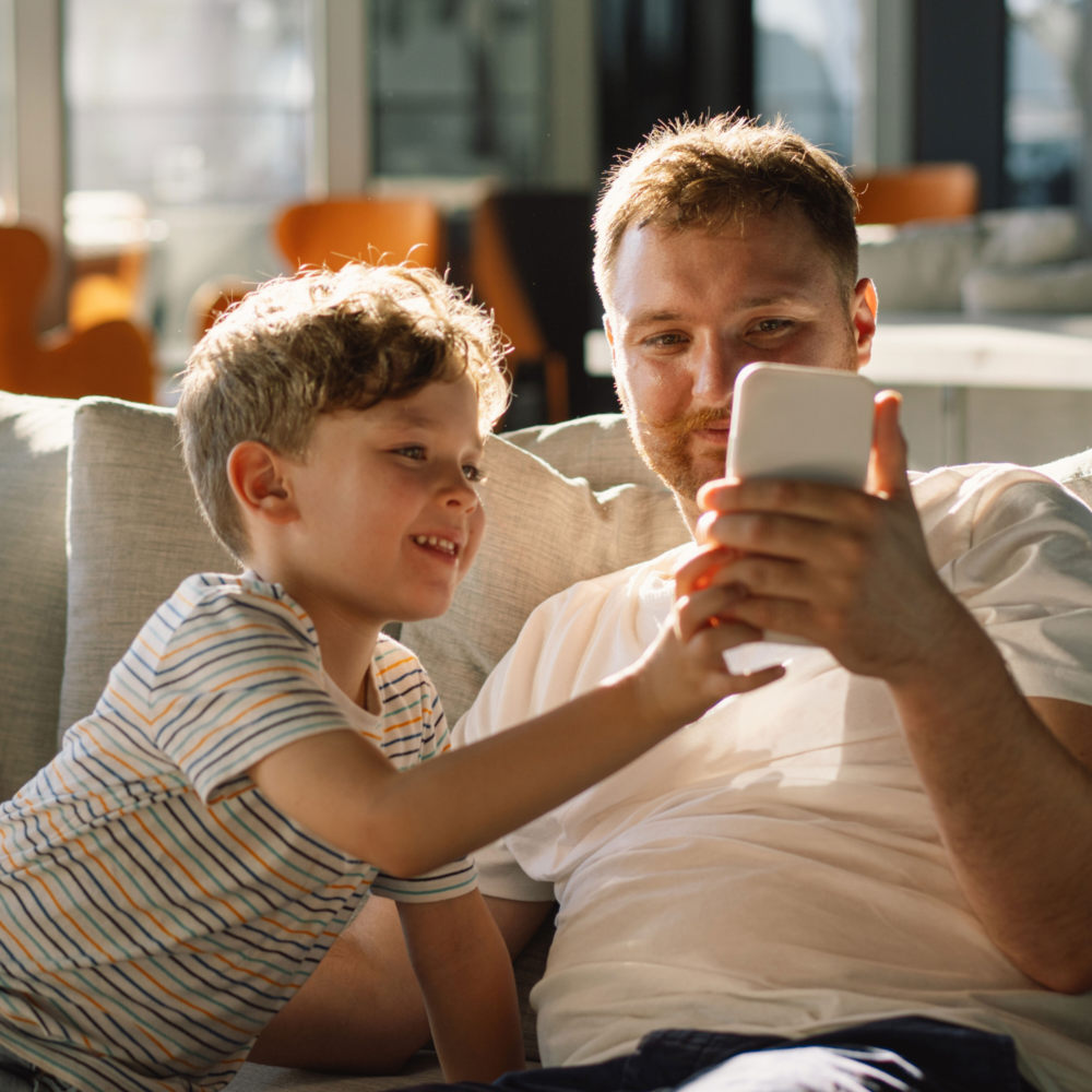 Ein Vater und sein kleiner Sohn sitzen eng beieinander auf einem hellen Sofa und schauen gemeinsam lachend auf ein Smartphone, das der Vater hält. Die Szene ist in warmes, natürliches Sonnenlicht getaucht. Das Bild illustriert begleitete Mediennutzung, das Teilen von digitalen Inhalten als verbindendes Element und eine positive Vater-Kind-Interaktion. Es symbolisiert digitale Nähe, die Bedeutung von gemeinsamem Erleben in der Medienpädagogik und die Beratung des Psychosozialen Gesundheitszentrums zur Förderung einer gesunden und gemeinschaftlichen Nutzung digitaler Medien in der Familie.