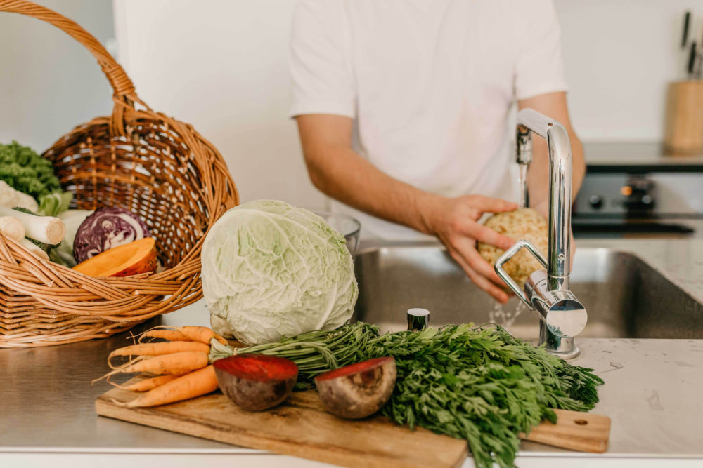 Eine Person wäscht Gemüse, daneben stehen ein Brett und ein Korb mit weiterem gesundem Gemüse.