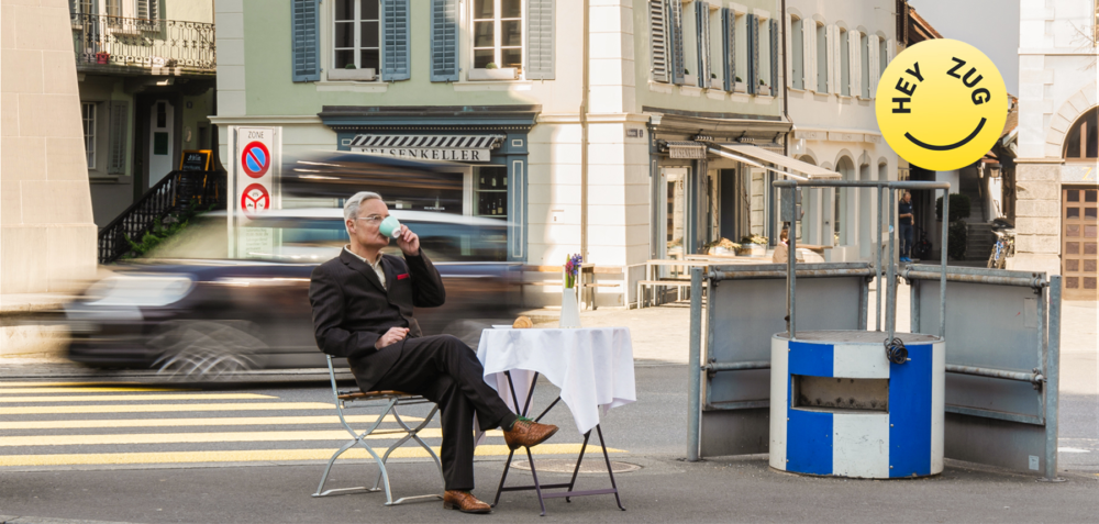 Eine Person sietzt auf einem Stuhl mit gedektem Tisch direkt an einer Strasse. Dahinter fährt ein Auto durch.