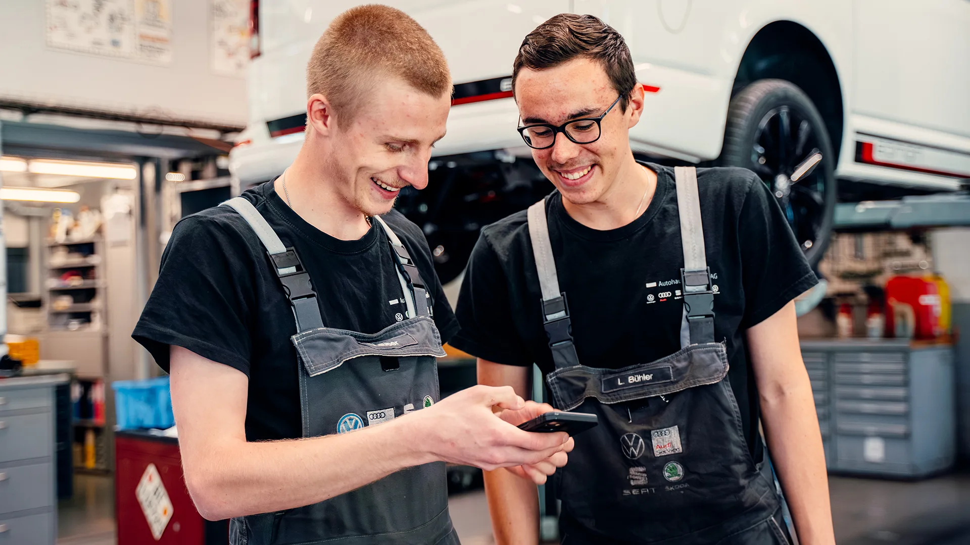 Zwei Mitarbeitende der Firma Autohaus von Känel stehen vor einem aufgebockten Auto und blicken lächelnd auf ein Smartphone.