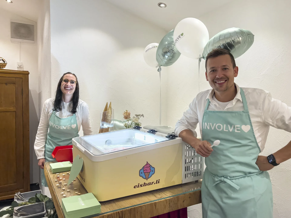 Zwei Mitarbeitende der Involve stehen lächelnd vor einer Eismaschine, aufgenommen beim Glace-Event Familienhilfe Lichtenstein