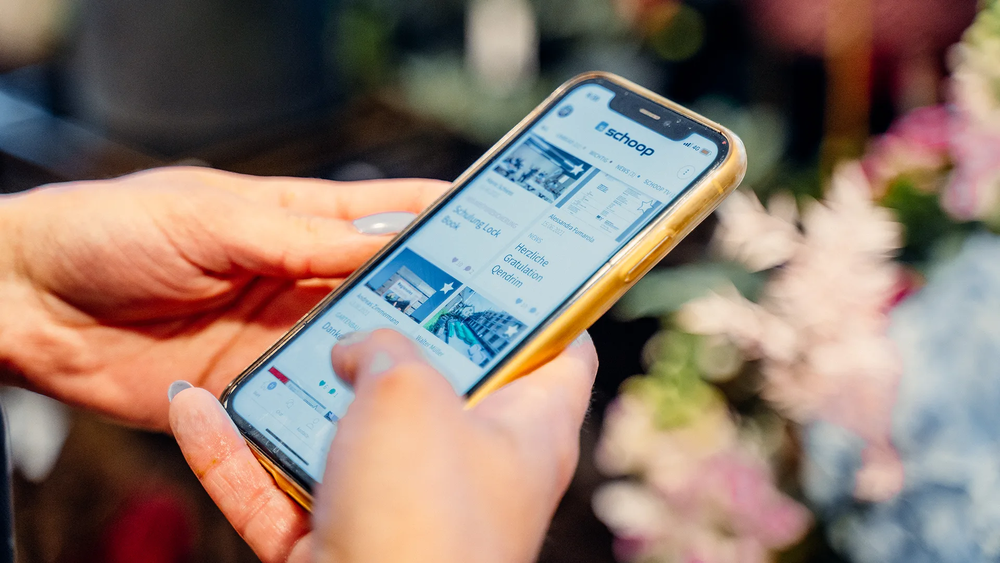 Smartphone wird in den Händen gehalten; auf dem Bildschirm ist die geöffnete Mitarbeitenden-App der Firma Schoop zu sehen.