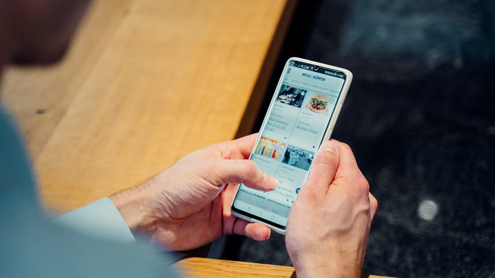 Smartphone wird mit beiden Händen gehalten; auf dem Bildschirm ist die geöffnete Mitarbeitenden-App der Firma Wild & Küpfer zu sehen