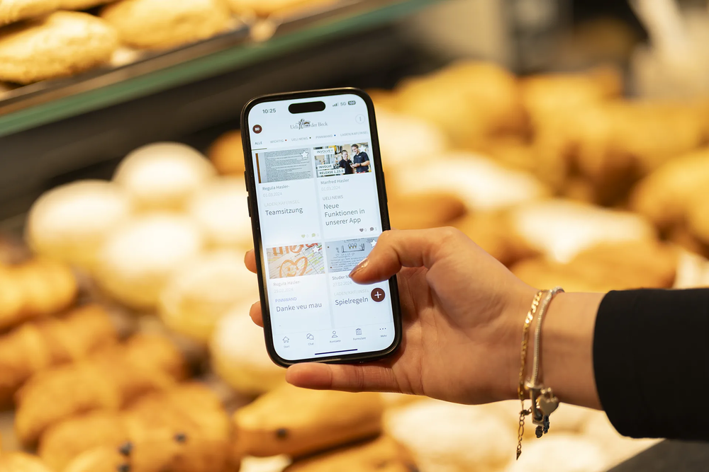 Frauenhand hält ein Smartphone mit geöffneter Ueli Beck Mitarbeitenden-App; im Hintergrund sind Backwaren zu sehen, aufgenommen 2024