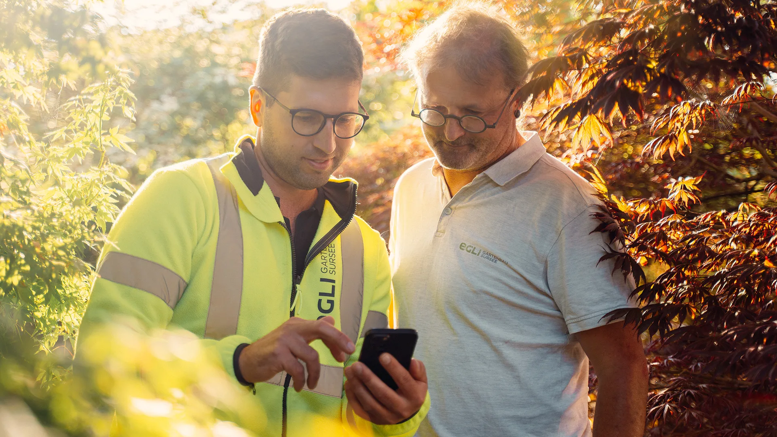 Zwei Mitarbeiter der Firma Egli Gartenbau nutzen die Mitarbeiter-App von Involve zur internen Kommunikation. Sie stehen zwischen Bäumen, aufgenommen 2023