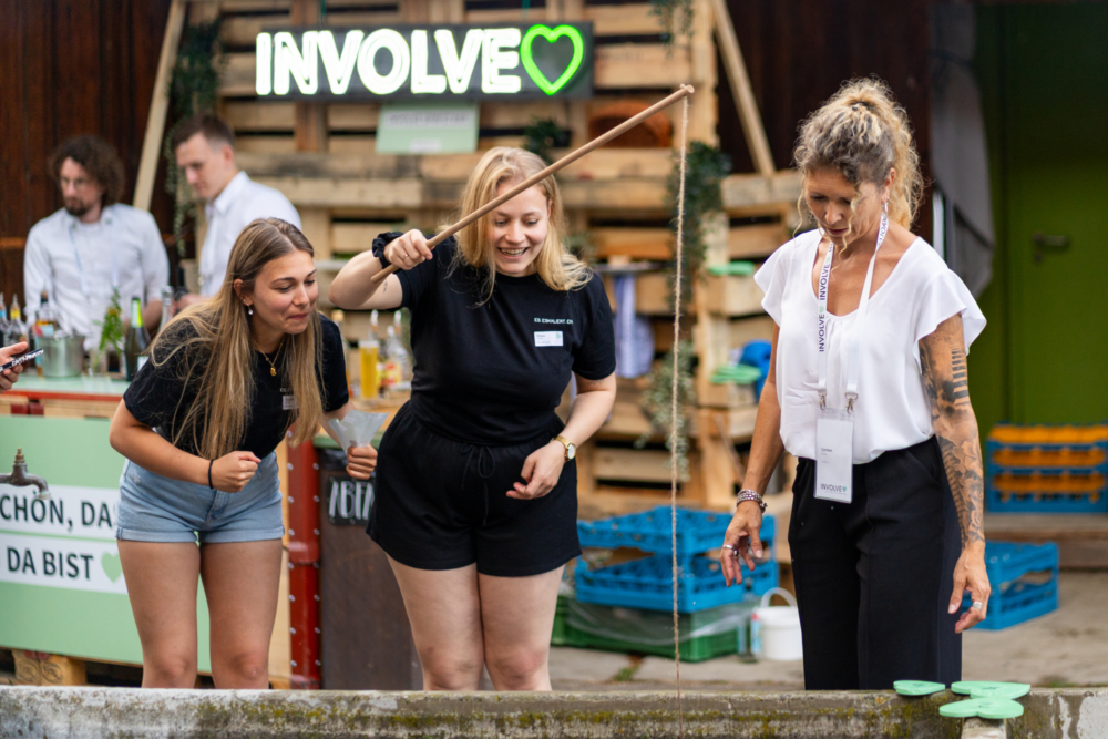 Zwei Kundinnen an der Involve Night 2025 im Burgrain stehen gemeinsam mit einer Involve-Mitarbeiterin vor einem Brunnen. Eine Kundin hält lachend eine Angel in der Hand.