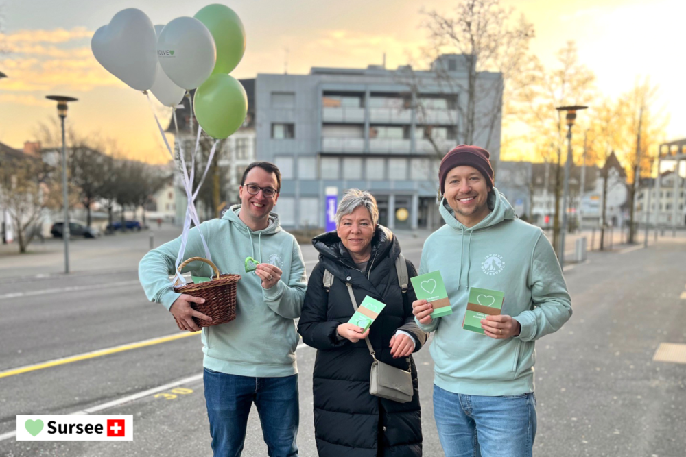 Zwei Mitarbeitende der Involve AG stehen mit einer Passantin auf dem Trottoir in Sursee; der linke hält einen mit Ballonen geschmückten Korb, alle lächeln in die Kamera.