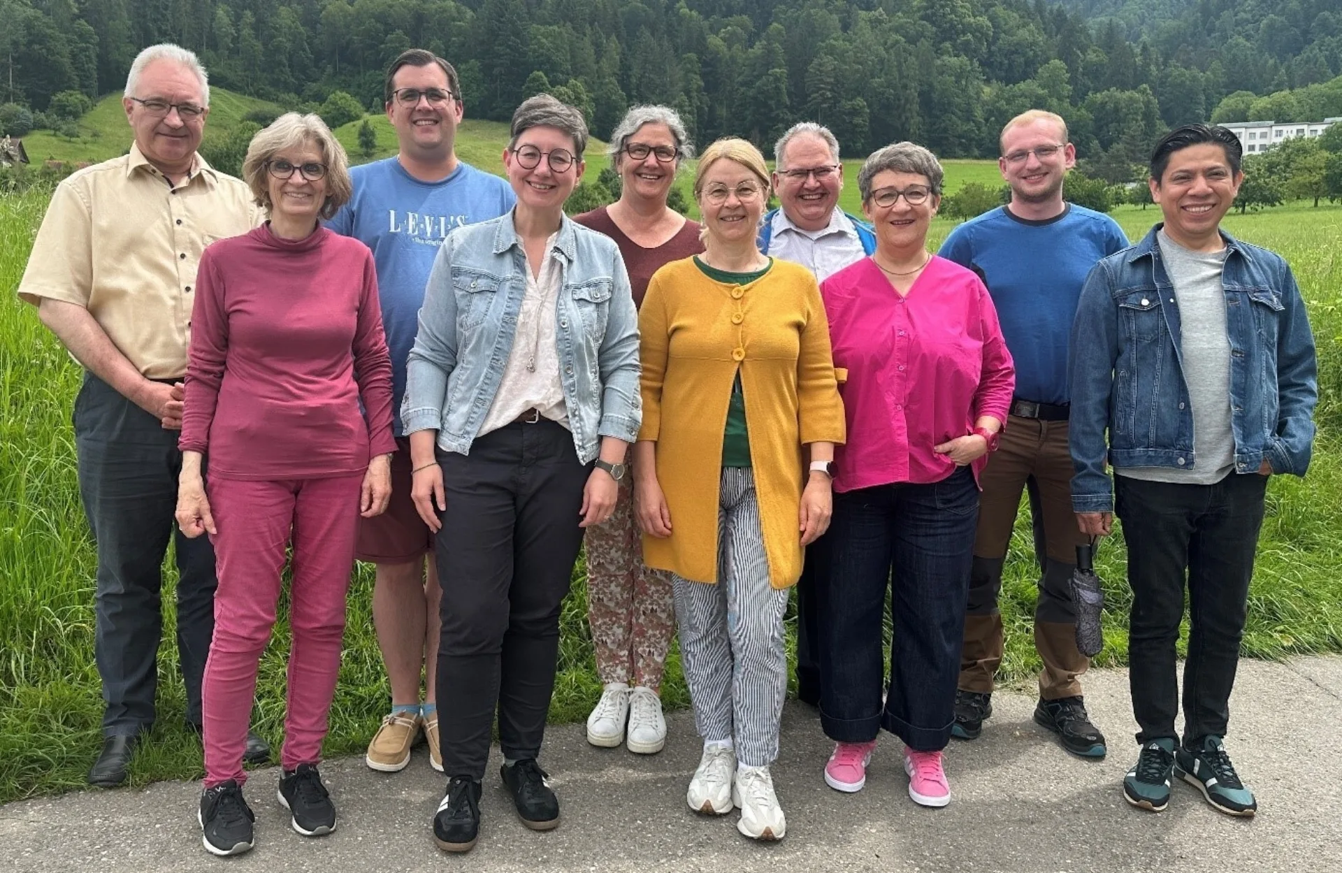 10 Personen, die das Pfarreiteam Bruder Klaus bilden, lachend vor einer grünen, hügeligen Wiese und Bäume im Hintergrund