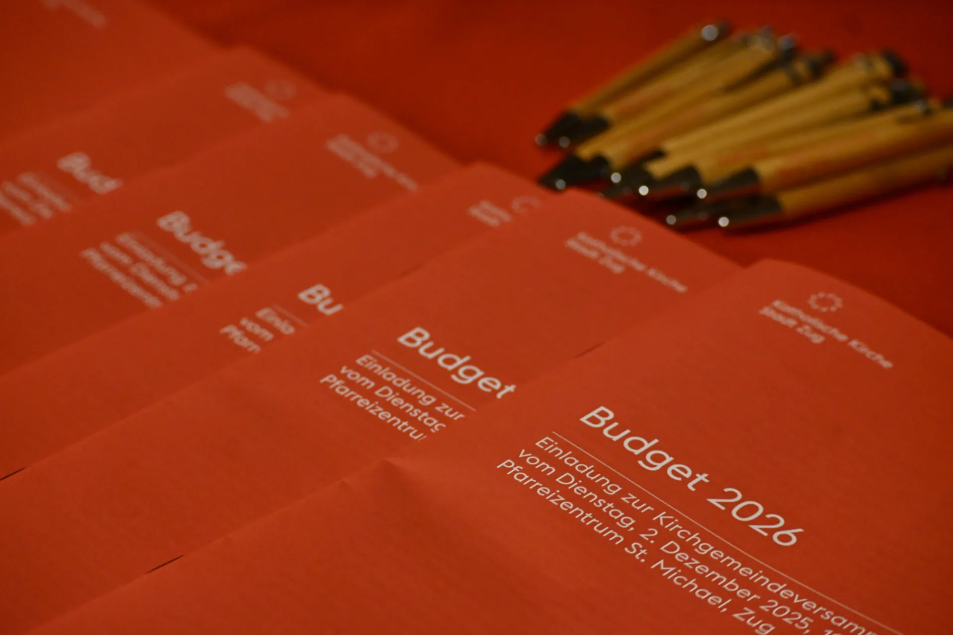 Rote Broschüren mit dem Titel "Budget 2026" liegen aufgereiht auf einem Tisch. Oben liegen Kugelschreiber.