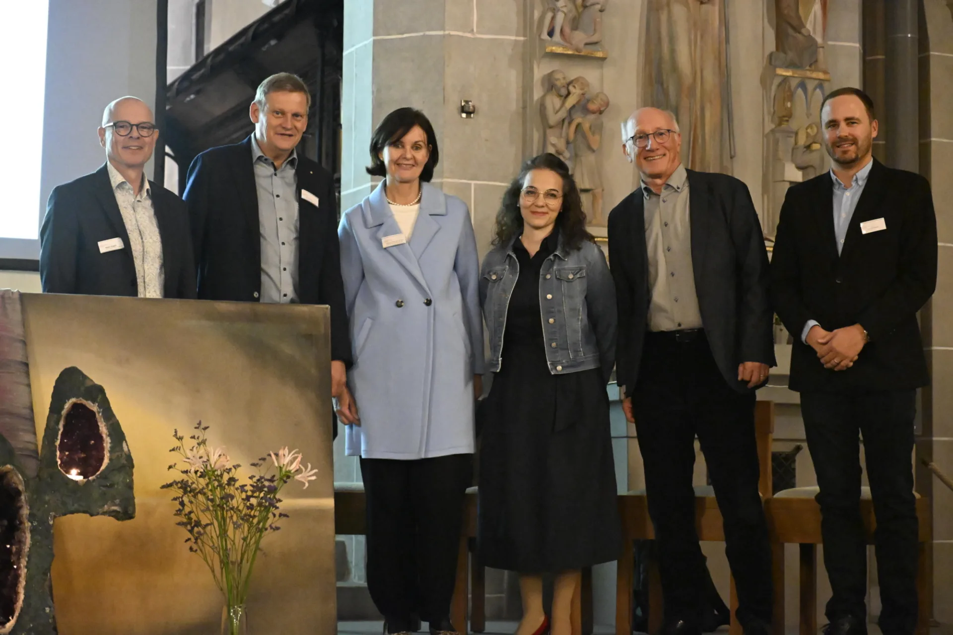 Sechs Personen, vier Männer im dunklen Anzug und in der Mitte zwei Frauen, stehen in einer Kirche. Vor den beiden Männern links ein bild in Goldtönen gehalten.