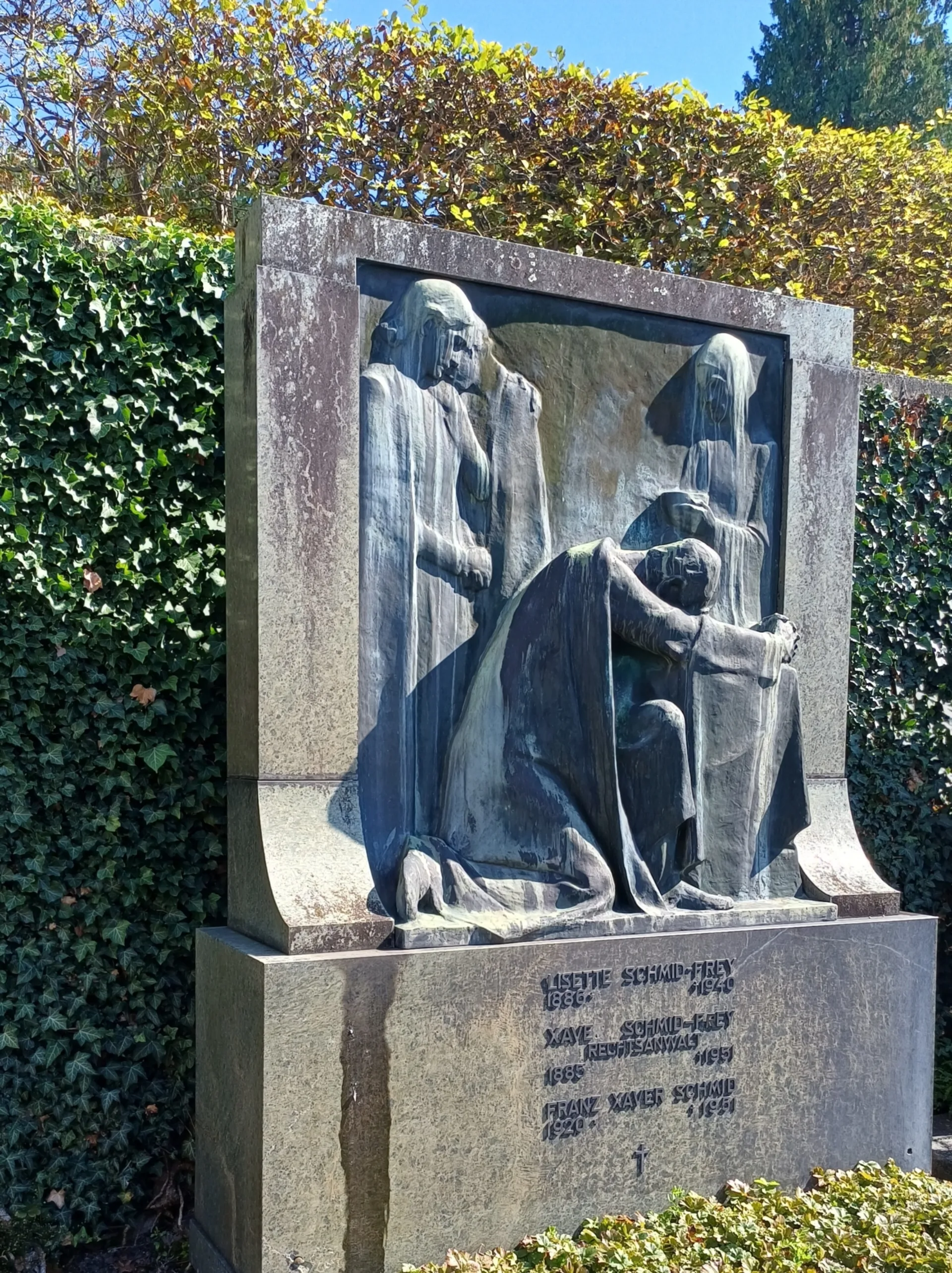 Ein Bronzerelief des Bildhauers Andreas Kögler stellt eine Gruppe Trauernder dar. Eine Person kniet, drei Personen in Hintergrund. Weiche Konturen, fliessende Gewänder. Ein Bronzerelief des Bildhauers Andreas Kögler stellt eine Gruppe Trauernder dar. Eine Person kniet, drei Personen in Hintergrund. Weiche Konturen, fliessende Gewänder.