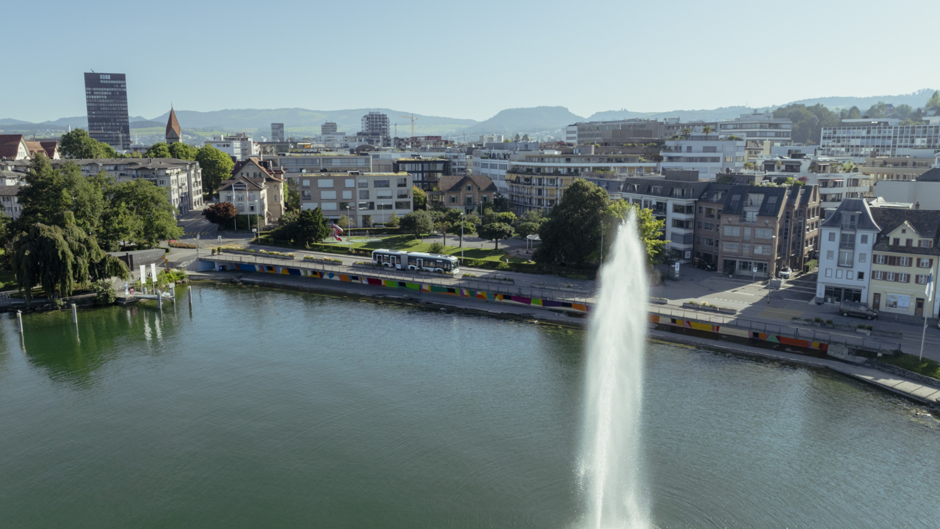 Blick auf die Häuser von Zug mit Zugersee, Springbrunnen und Bus auf der Strasse.